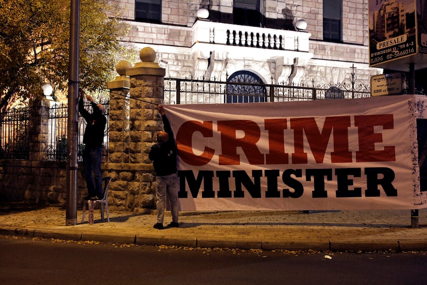 You view protesters putting up a banner that reads 'Crime Minister' in on the gates of a large residence at night.