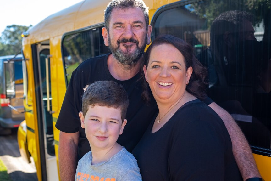 A mum, dad and son stand in front of a yellow school bus.