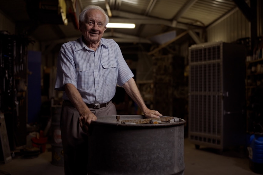 An elderly man stands in a shed that is set up as a workshop with plenty of tools and cupboards. He is leaning on a steel barrel