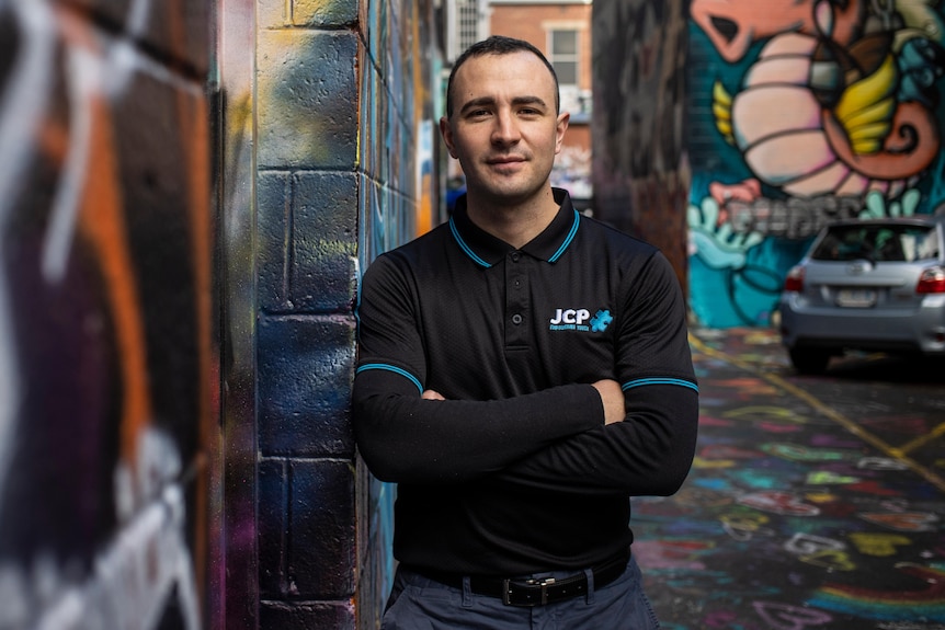 A man crosses his arms and leans against a graffitied wall.