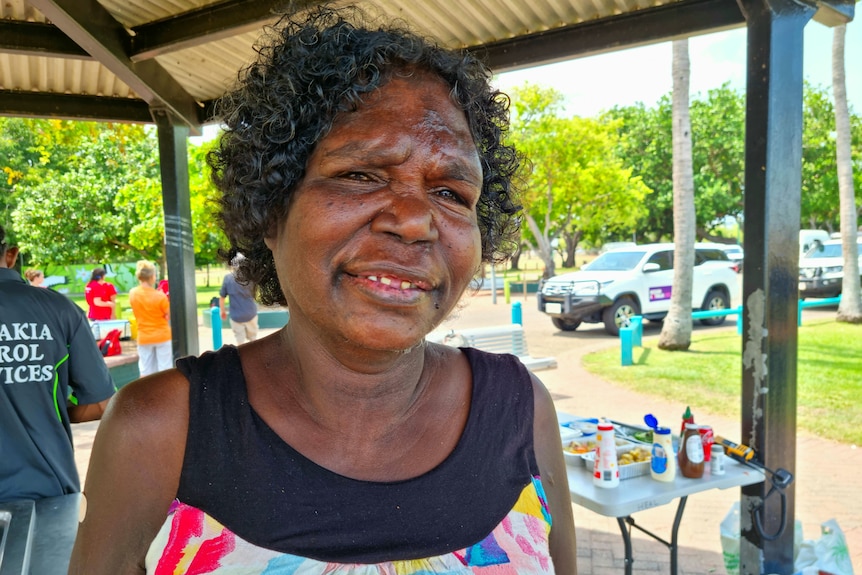 A smiling woman in a colourful dress under a shade structure, with greenery in the background. 