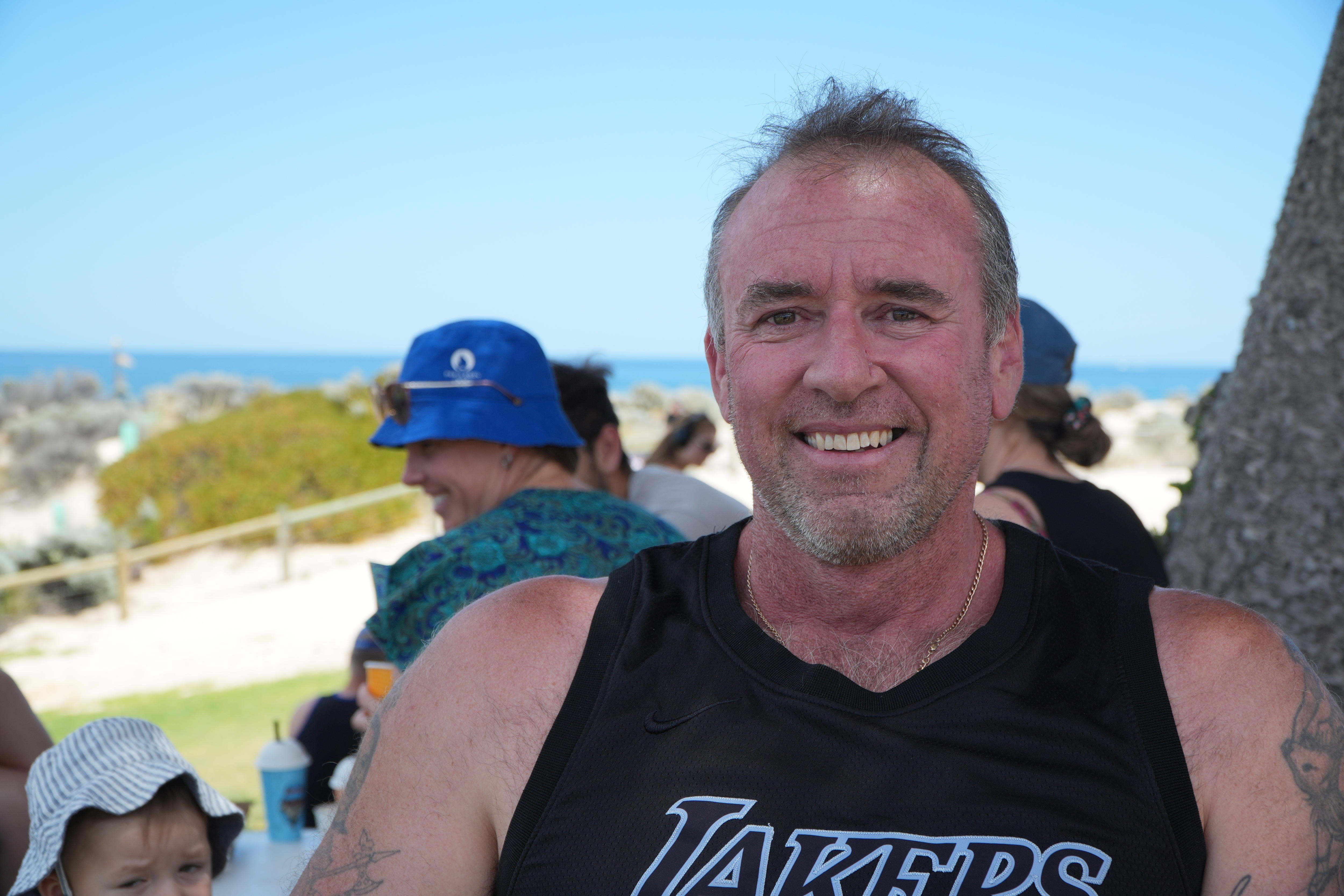 A middle-aged man in a tank top grins beside a beach.