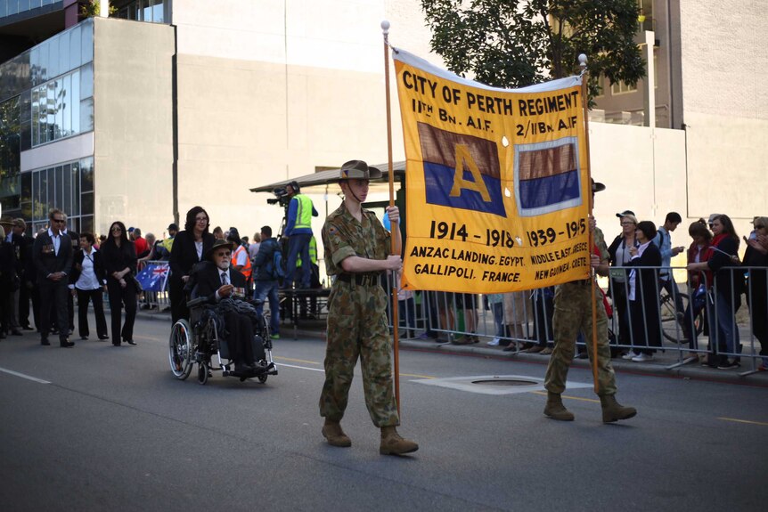 Two soldiers carrying a yellow banner march in front of a man in a wheelchair.