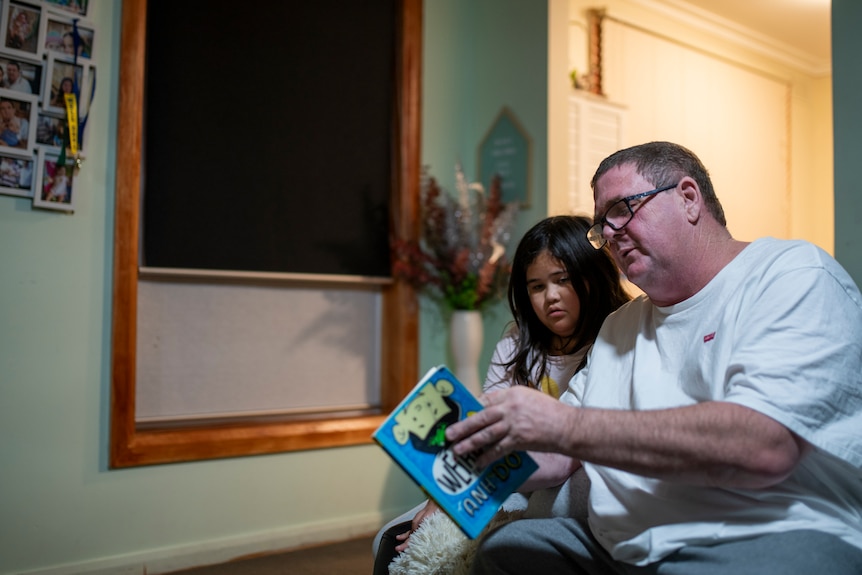 A man in glasses and a white t-shirt holds a book out as a younger girl reads aloud and leans into him in their living room.