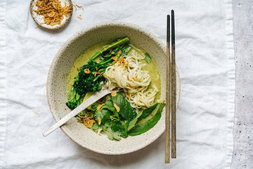 Bowl of Thai green curry noodle soup topped with basil and coriander, fried shallots to serve on the side.