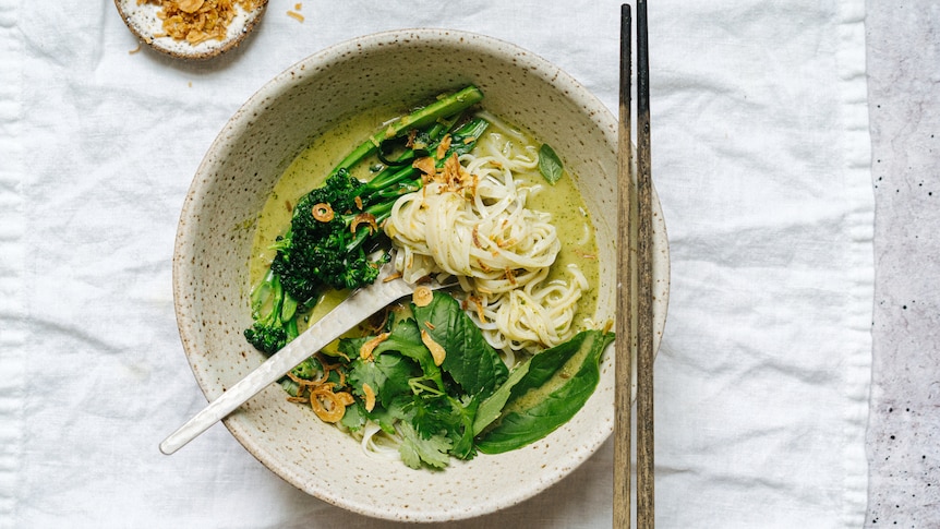 Bowl of Thai green curry noodle soup topped with basil and coriander, fried shallots to serve on the side.