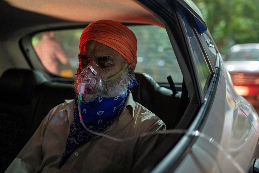 An elderly man in a turban receives oxygen support in the back of a car