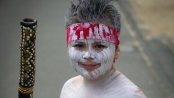 An Indigenous boy wearing white body paint and a red headband stands smiling and holding a didgeridoo.