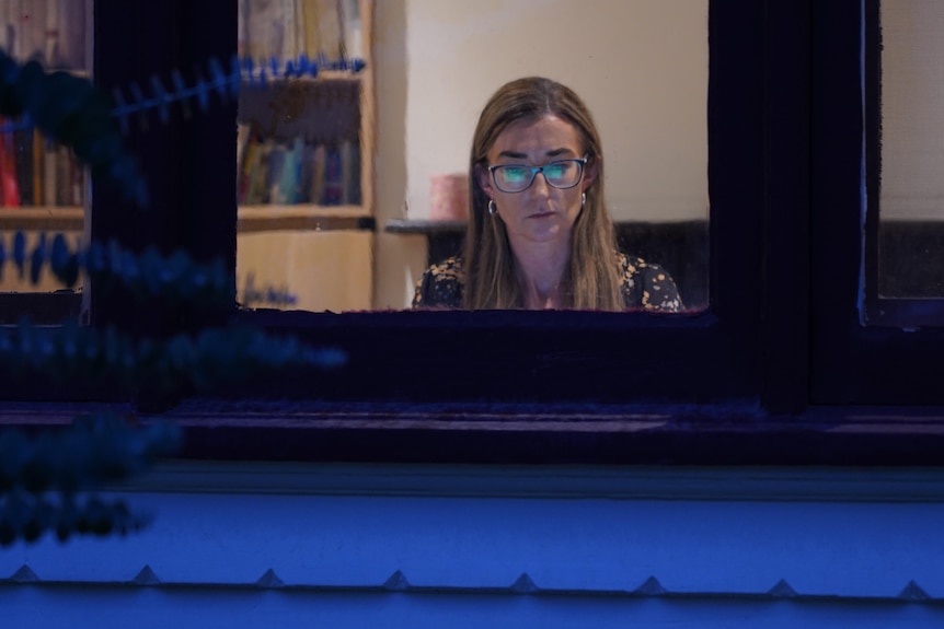 A woman with dark blonde hair and glasses sits at computer 