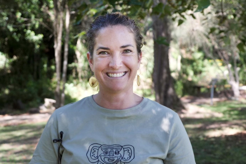 A woman in a khaki T-shirt smiling warmly with a bush backdrop of dappled light out of focus behind her.