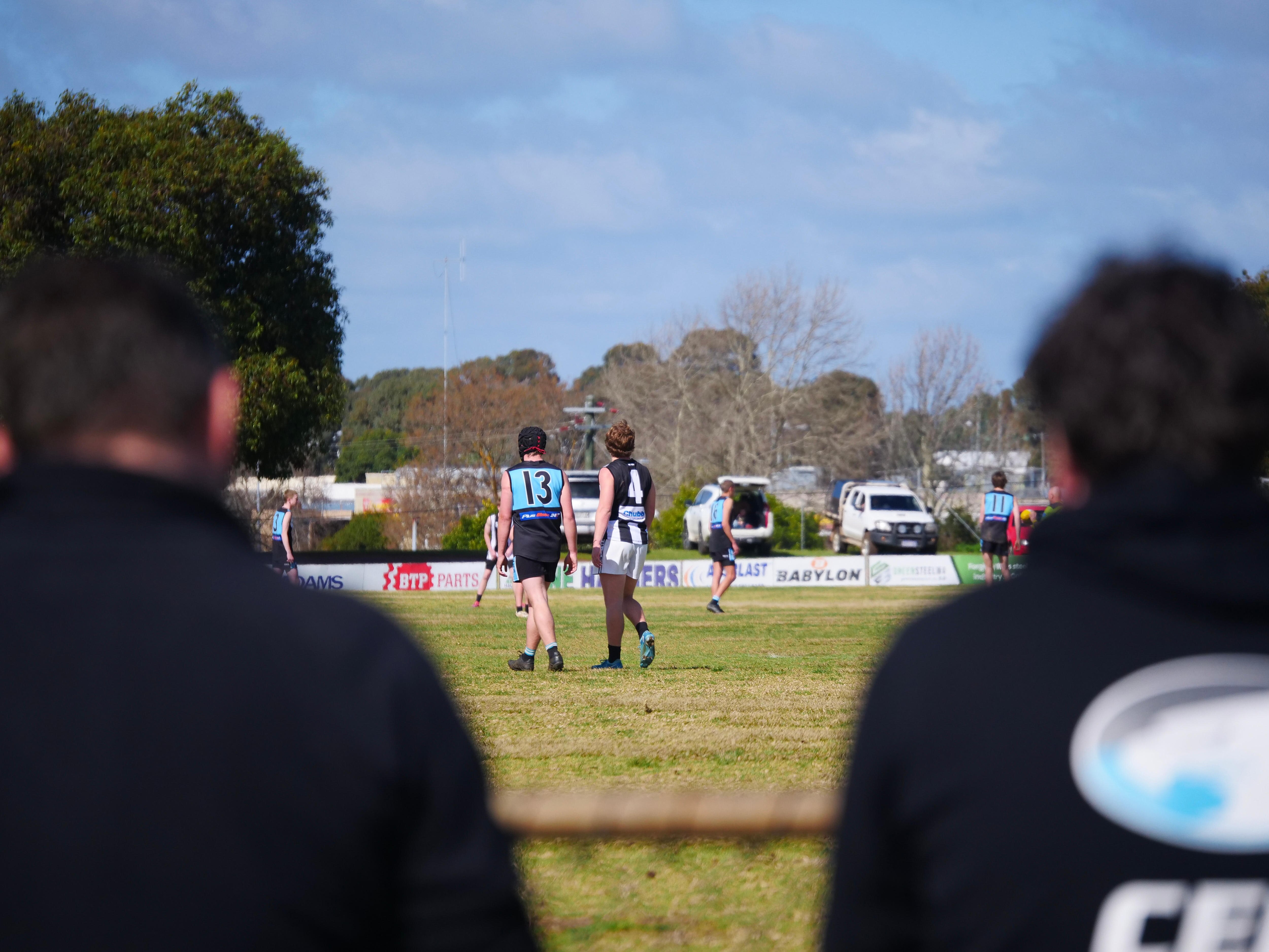View from behind two men watching local football match on grass oval