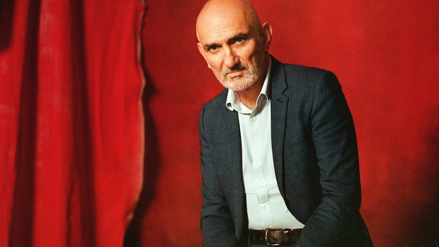 A stubble-faced Paul Kelly wears a suit and sits in front of a red curtain.