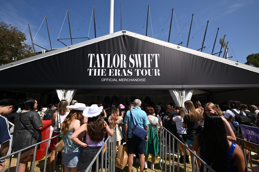 Fans in costume line in a queue to buy merchandise with a black 'Taylor Swift The Eras Tour' sign