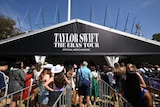 Fans in costume line in a queue to buy merchandise with a black 'Taylor Swift The Eras Tour' sign