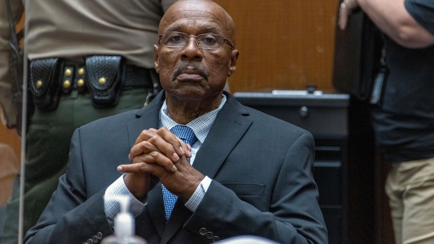 A bespectacled black man dressed in a suit claps his hands together as he sits in a courtroom