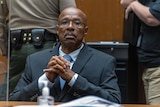 A bespectacled black man dressed in a suit claps his hands together as he sits in a courtroom