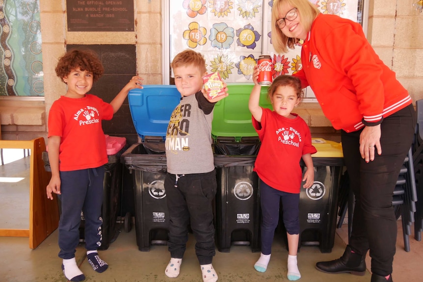 Three preschool students and their teacher posing with recyclable items in front of three small recycling waste bins
