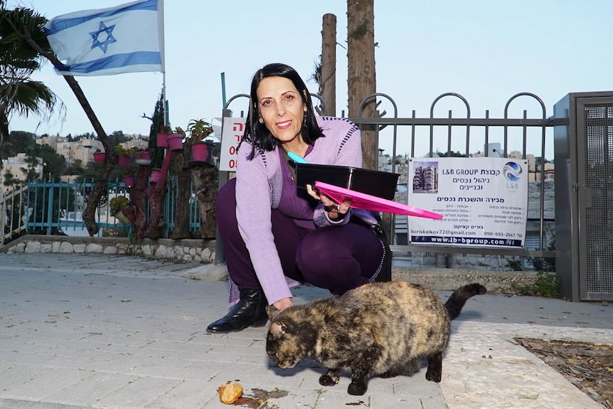 Ilana Ben Joya crouches down next to a fluffy cat in front of a plastic container with food on an outdoor walkway