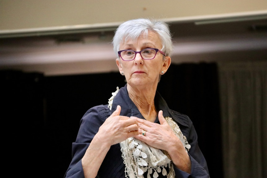 A woman with grey hair and purple classes, wearing a scarf and a black shirt, demonstrates a hand sign.