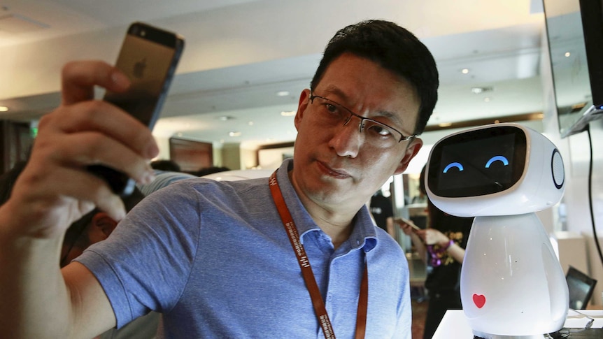 A visitor takes a selfie with Baidu's robot Xiaodu. It is a small white robot with curved eyes to make it look like it's smiling