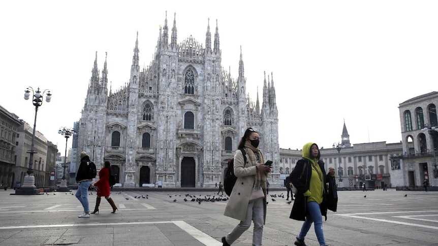 Four people stand in front of the square in front of Duomo gothic cathedral in Milan, one wearing a mask.
