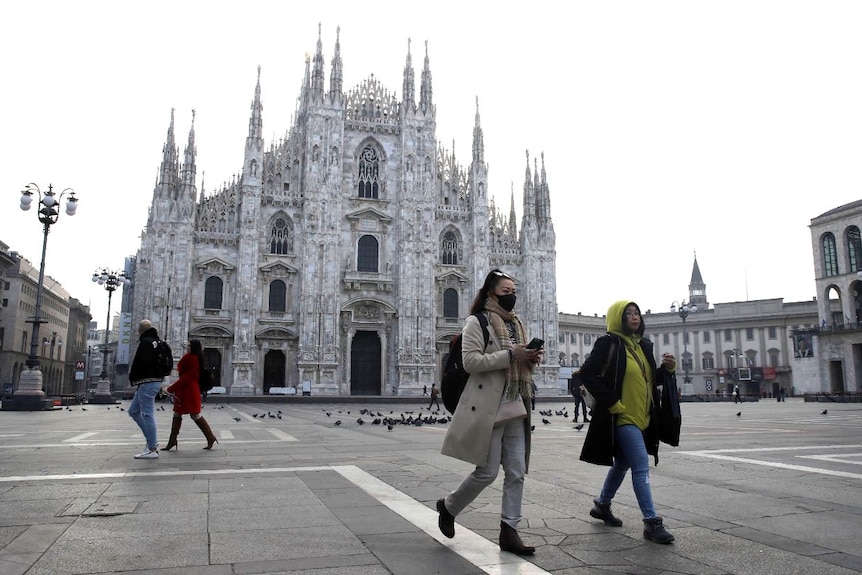 Four people stand in front of the square in front of Duomo gothic cathedral in Milan, one wearing a mask.