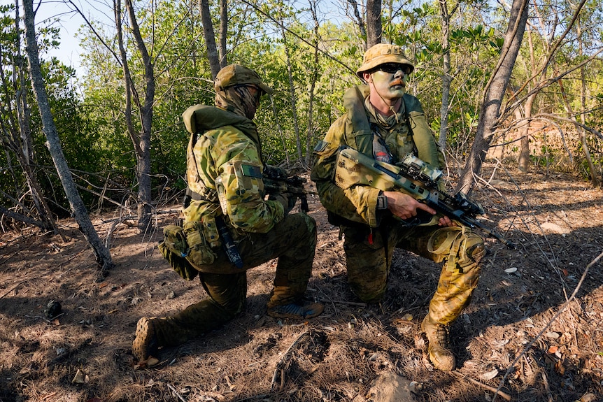 two men in army uniform kneeling in tropical bushland holding guns