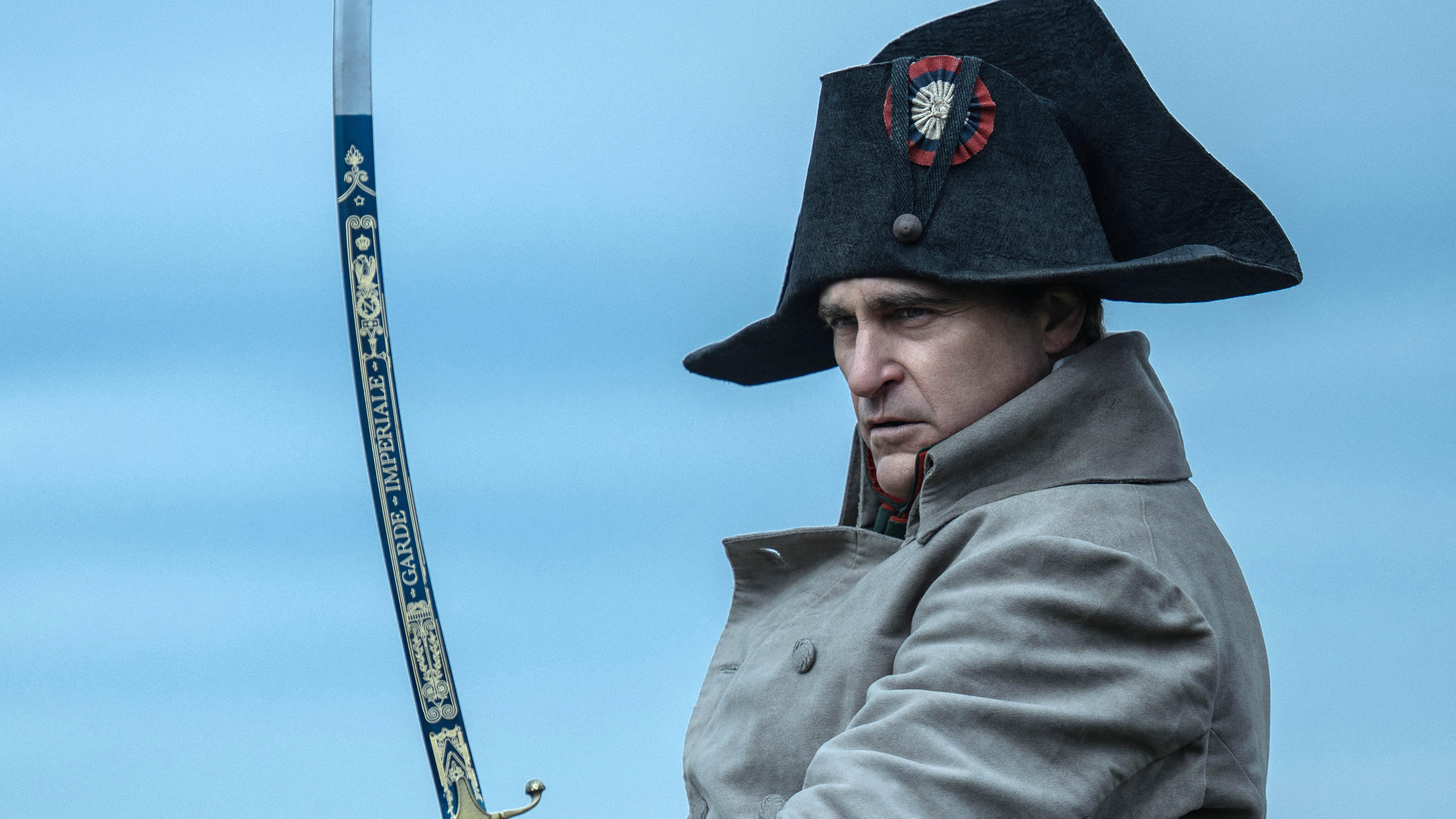 Joaquin Phoenix acting on screen as Napoleon Bonaparte in an 18th-century French naval uniform and large hat, wielding a sword.