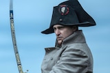 Joaquin Phoenix acting on screen as Napoleon Bonaparte in an 18th-century French naval uniform and large hat, wielding a sword.