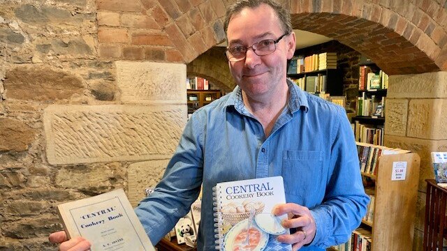 A man in a blue shirt and glasses holds two editions of a cook book.