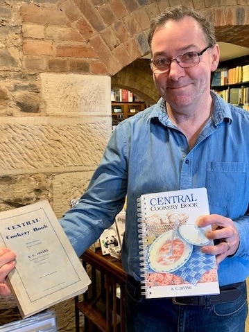 A man in a blue shirt and glasses holds two editions of a cook book.