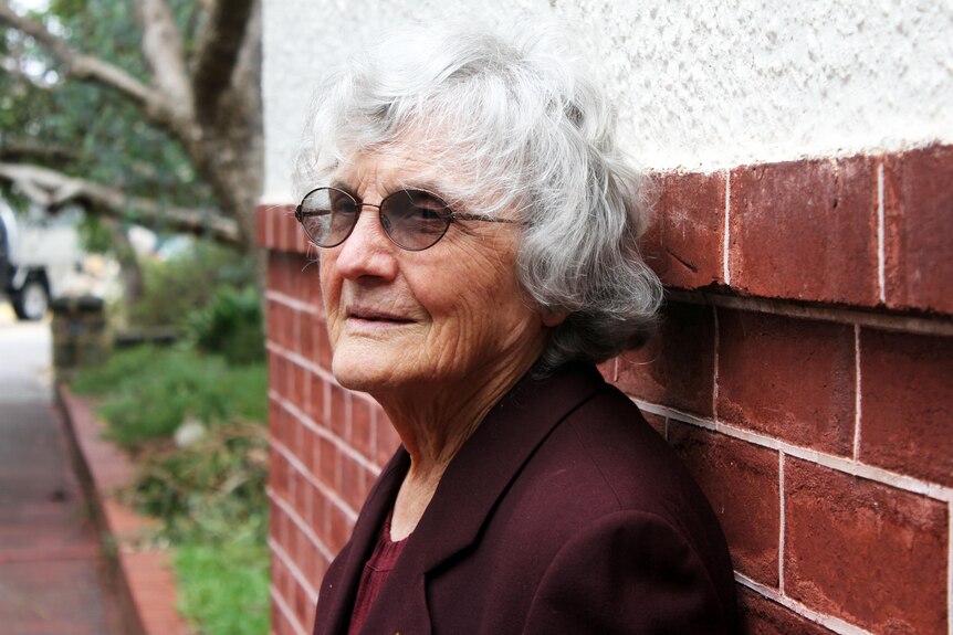 Silver-haired woman with glasses stands with her back to a red brick wall