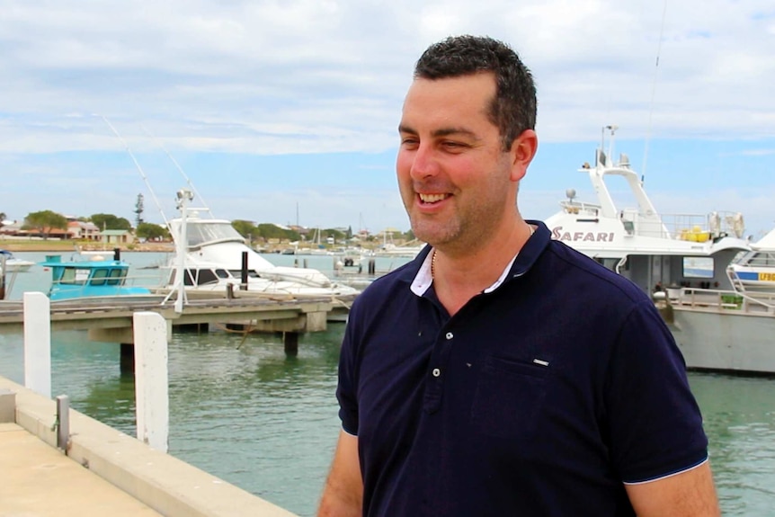 A man stands outside with lobster boats in the background