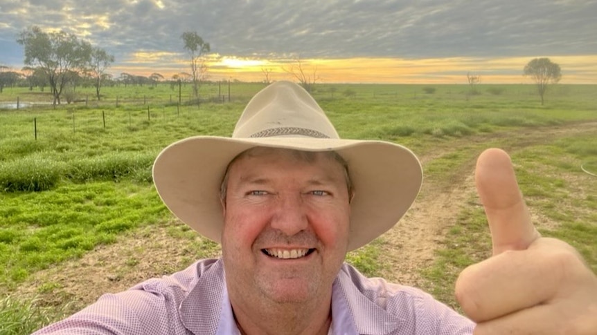 Selfie of  head of a smiling man wearing an Akubra hat giving a thumbs up with lush green landscape and sunset in the background