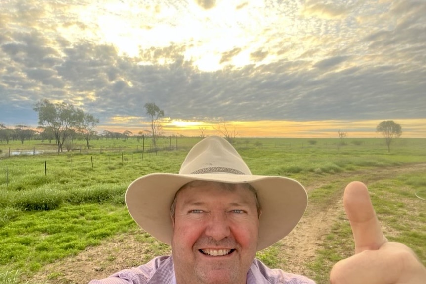 A man in an Akubra smiles and gives the thumbs up as he takes a sunset selfie in front of lush green paddocks.
