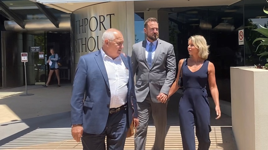 Man in grey suit walking out of court next to older man in blue suit and holding hands with blonde lady in blue jumpsuit