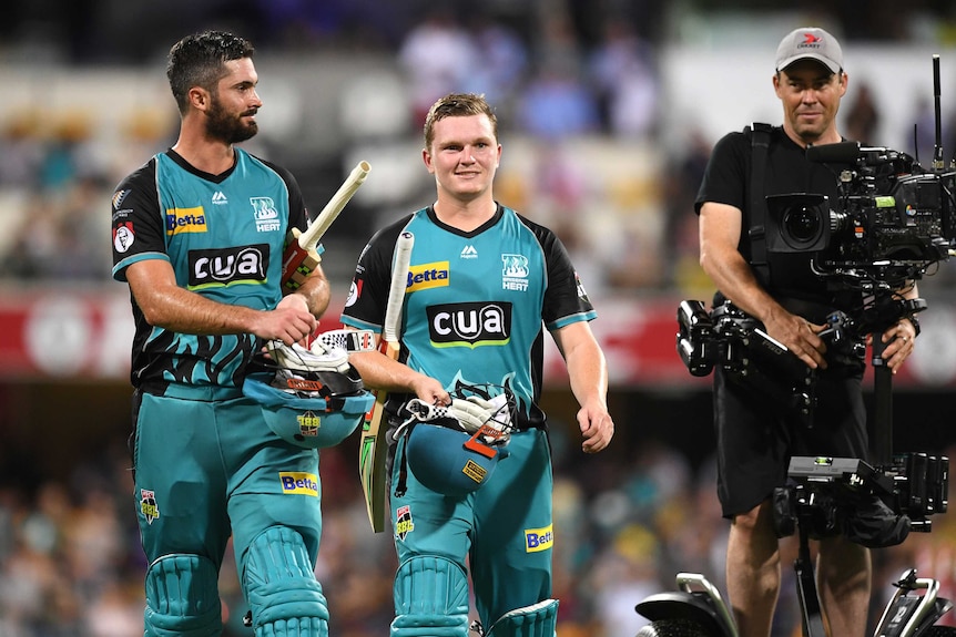 Cricket players walk off the field of play with a TV camera next to them.