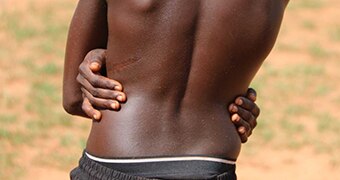 The back of a shirtless Aboriginal teenage boy wearing black pants and with his arms wrapped around his waist.