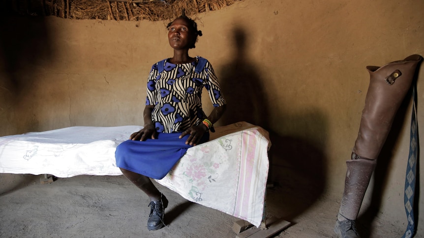 Winnie sits on a bed in a mud floor hut with her prosthetic leg beside her