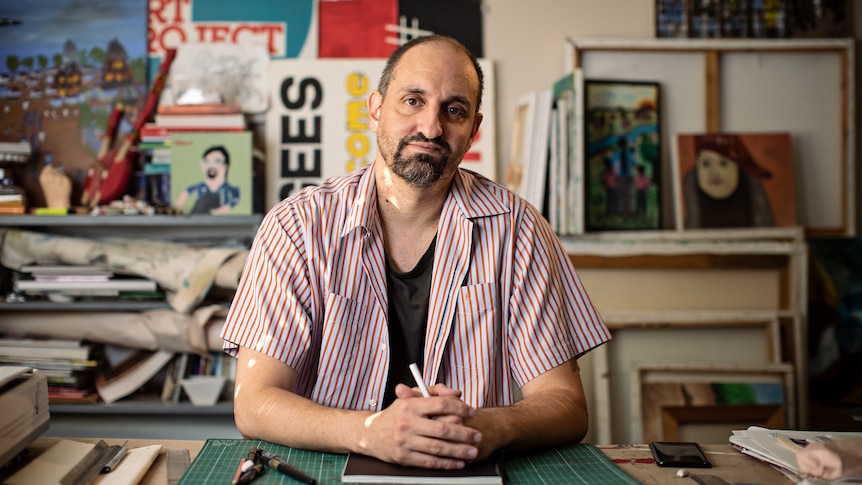 A man with a goatee sits behind a drawing desk, hands clasped around a marker, art lining the walls and shelves behind him