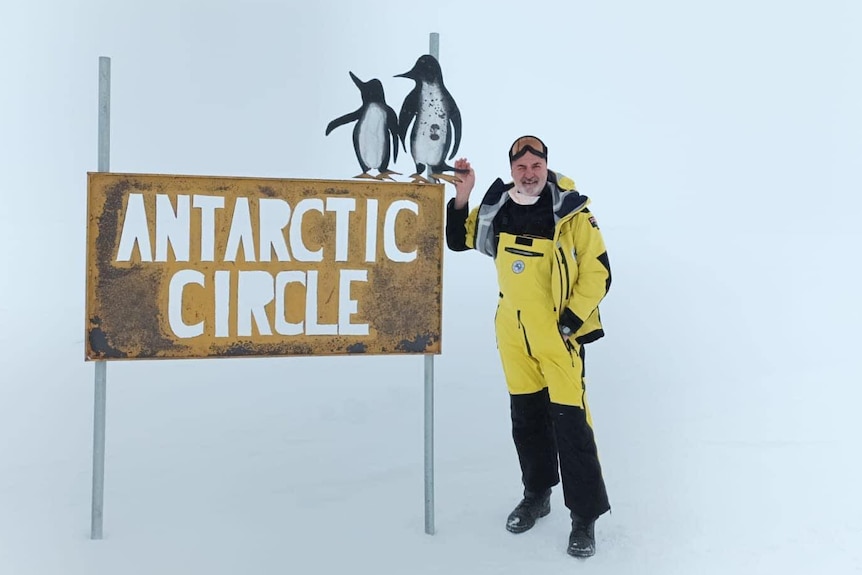 A man in a yellow snowsuit smiles as he stands in the snow next to a sign saying 'Antarctic Circle'.