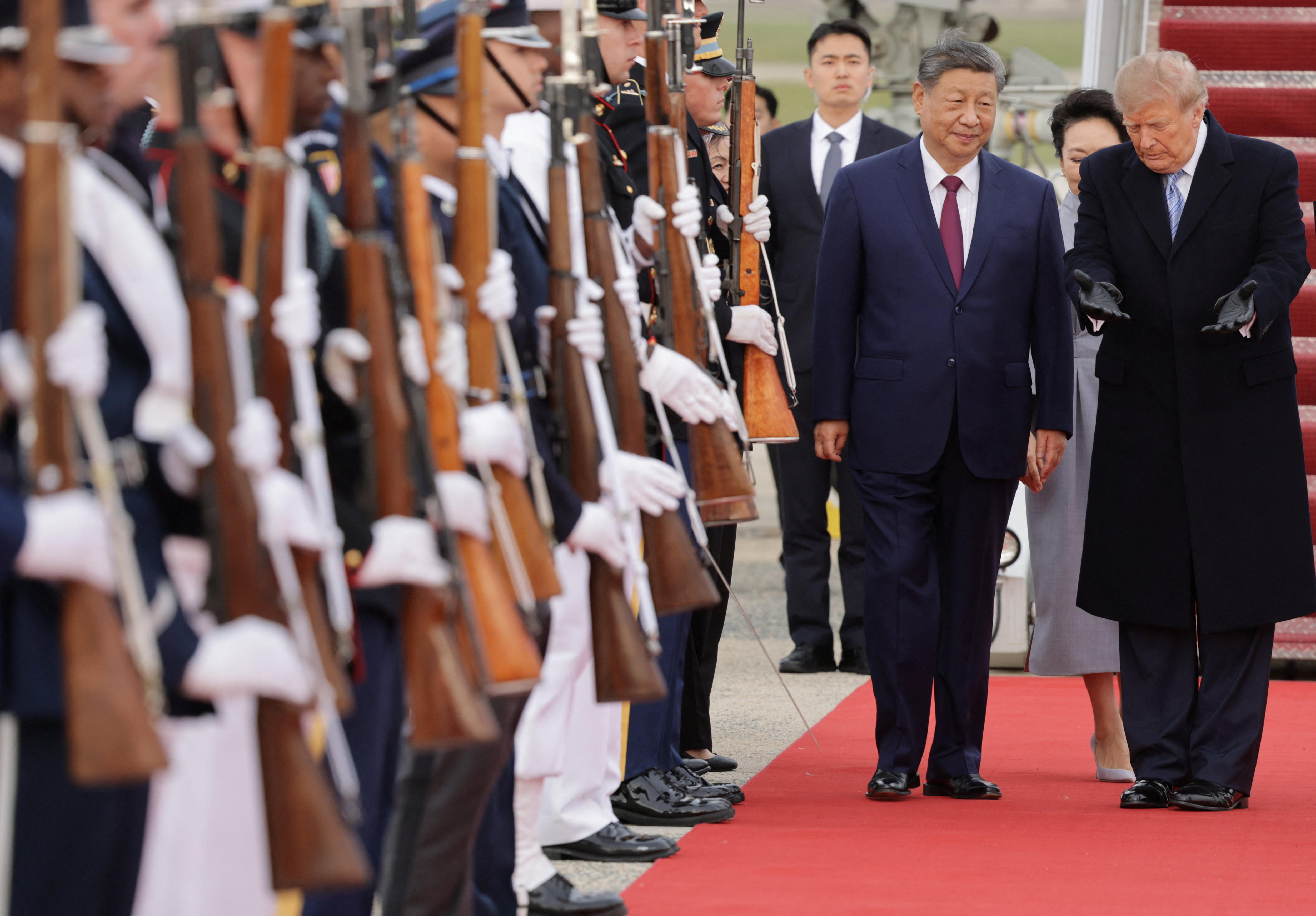 Two men stand next to each other with one gesturing to the other to walk past soldiers standing with guns
