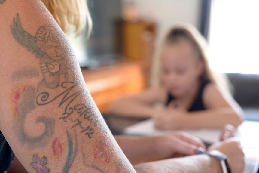 A woman's tattooed arm close up, a child sitting at a kitchen table blurred in the background