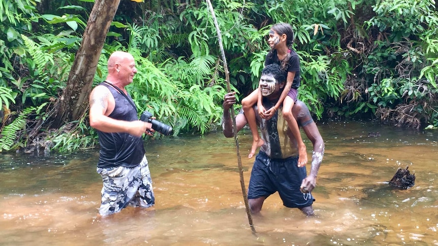 Man stands in shallow water of river with camera in hand waiting to photograph a man with a child on his shoulders