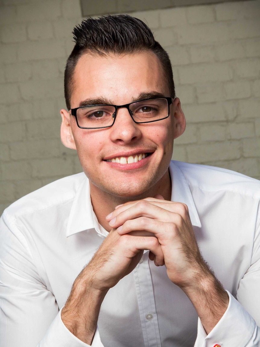 Alex Cowan sits with hands clasped in front of a brick wall.