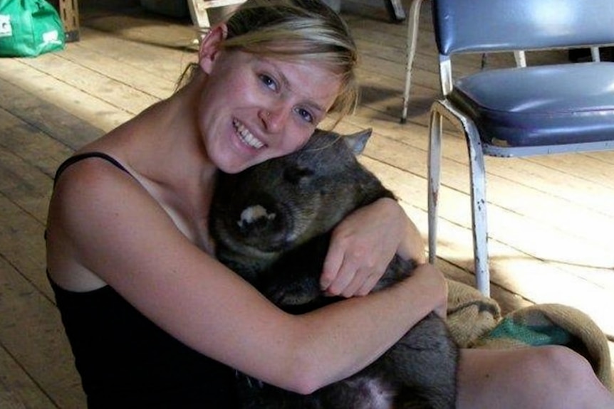 A young woman with blonde hair tied back sits on floor holding a large wombat