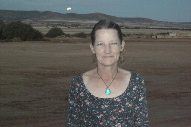 A middle-aged woman with paddocks, hills and the moon in the background.