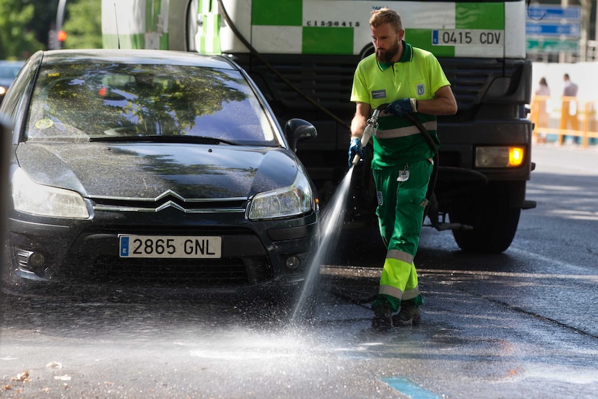 A Spanish municipal worker in bright and dark green hoses down an asphalt street next to a black Citroen.