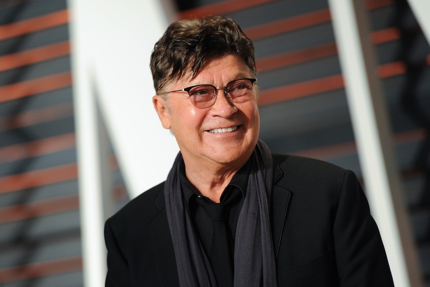 A man in a black shirt and scar, arrives at a premiere, smiling to photographers
