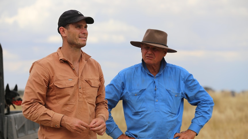 Two men stand outside in farming paddock, one wears a cap and another a hat.